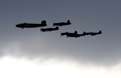 [Large, old warplane flying in front of four smaller planes that appear to be blacked out (so one sees the outline) because of the angle of the light and clouds.]